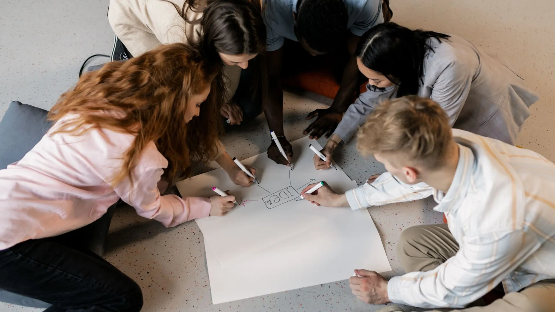 A multicultural team collaborates on an idea using markers and paper on the floor indoors.