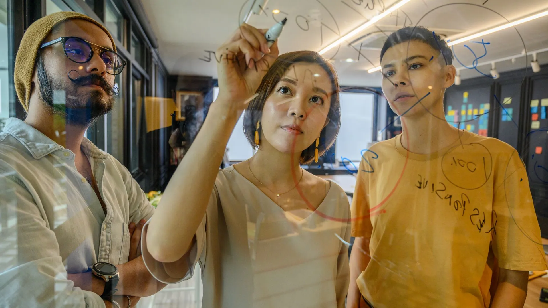 Three colleagues engaged in a strategy meeting, writing ideas on glass in a modern office.